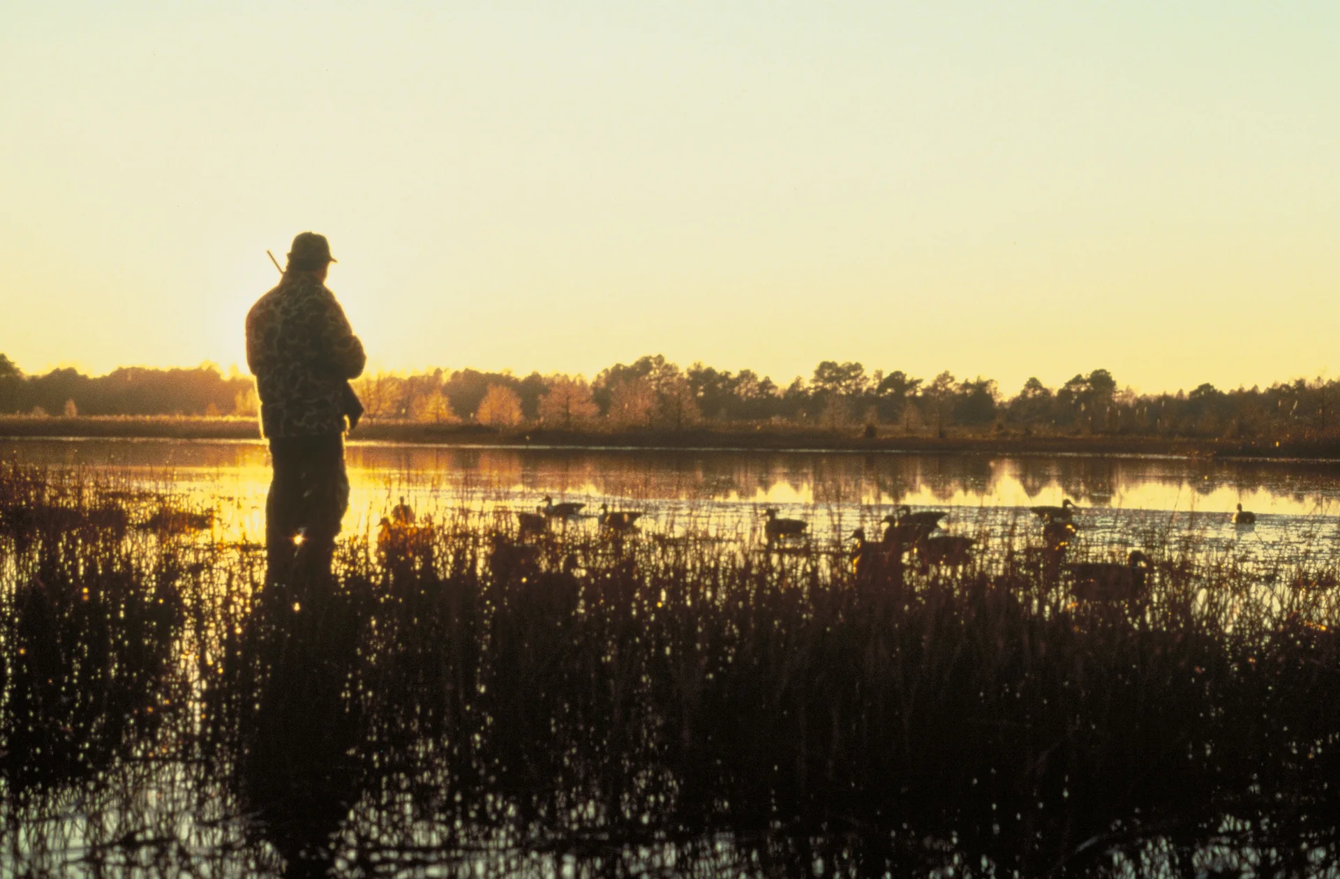 A silhouetted hunter stands beside decoys on quiet marsh water at sunrise.